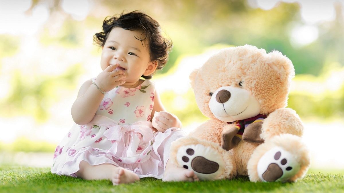 baby girl sitting on grass beside bear plush toy. Image courtesy of Singkham at Pexels