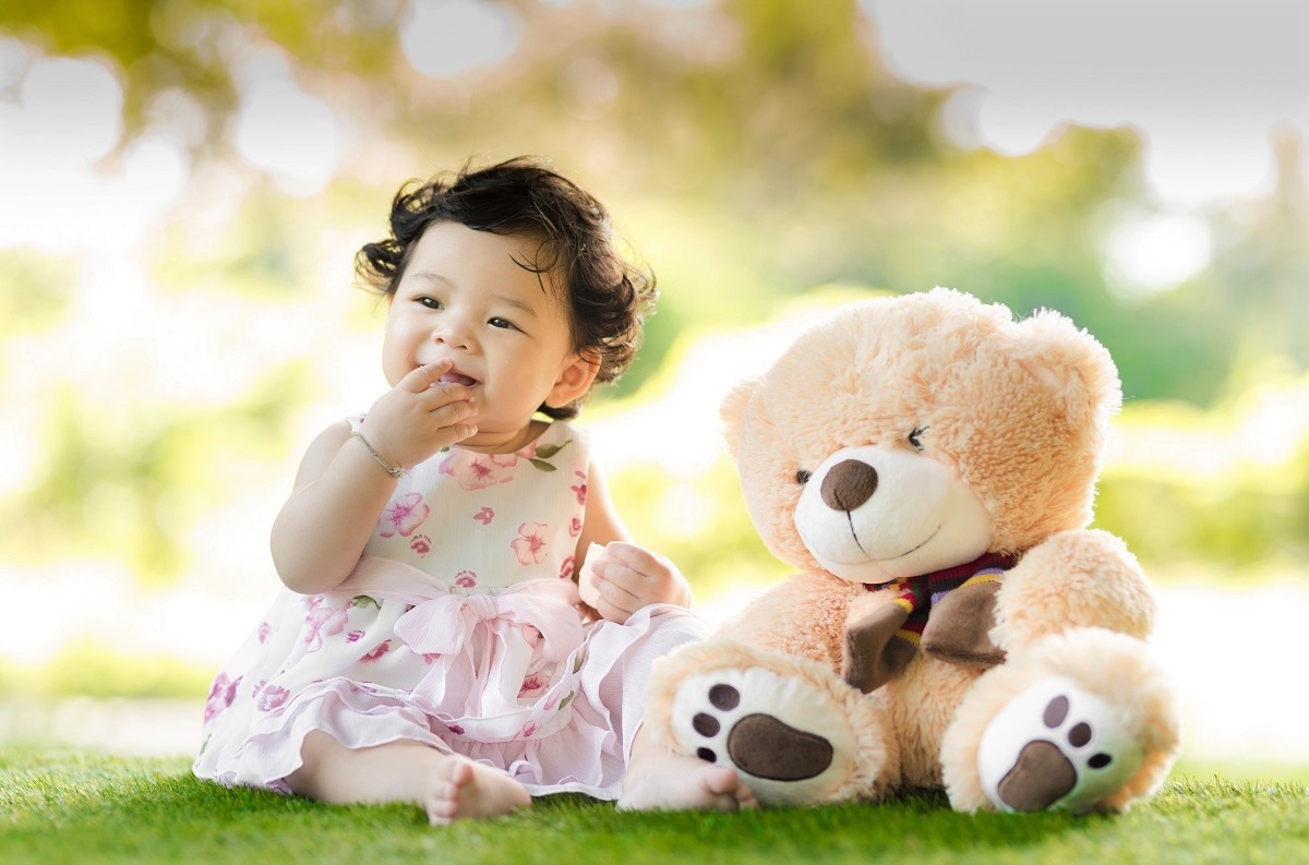 baby girl sitting on grass beside bear plush toy. Image courtesy of Singkham at Pexels