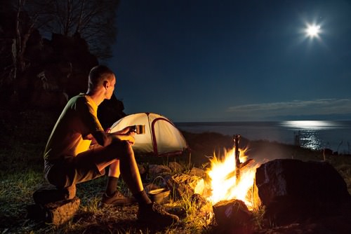 young man sitting at a campfire on ridge overlooking water