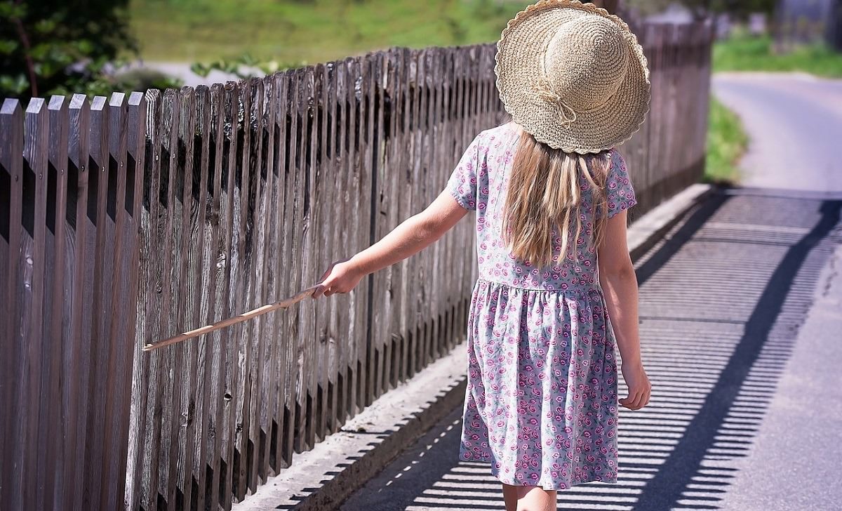 young girl dragging stick along wooden fence