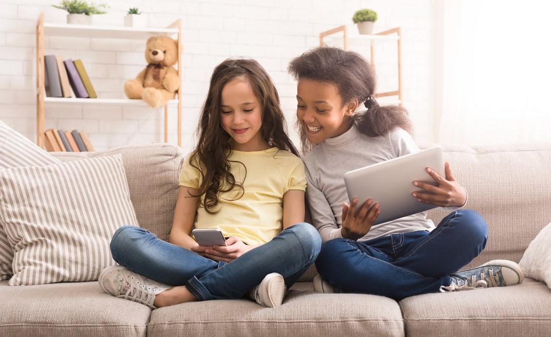 smiling girls sitting cross-legged on sofa with computer devices