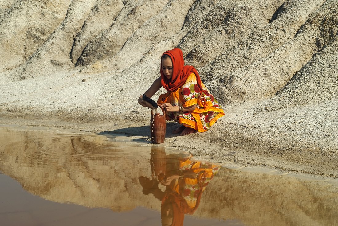 Woman in hijab fills a clay jar with brown water in arid region