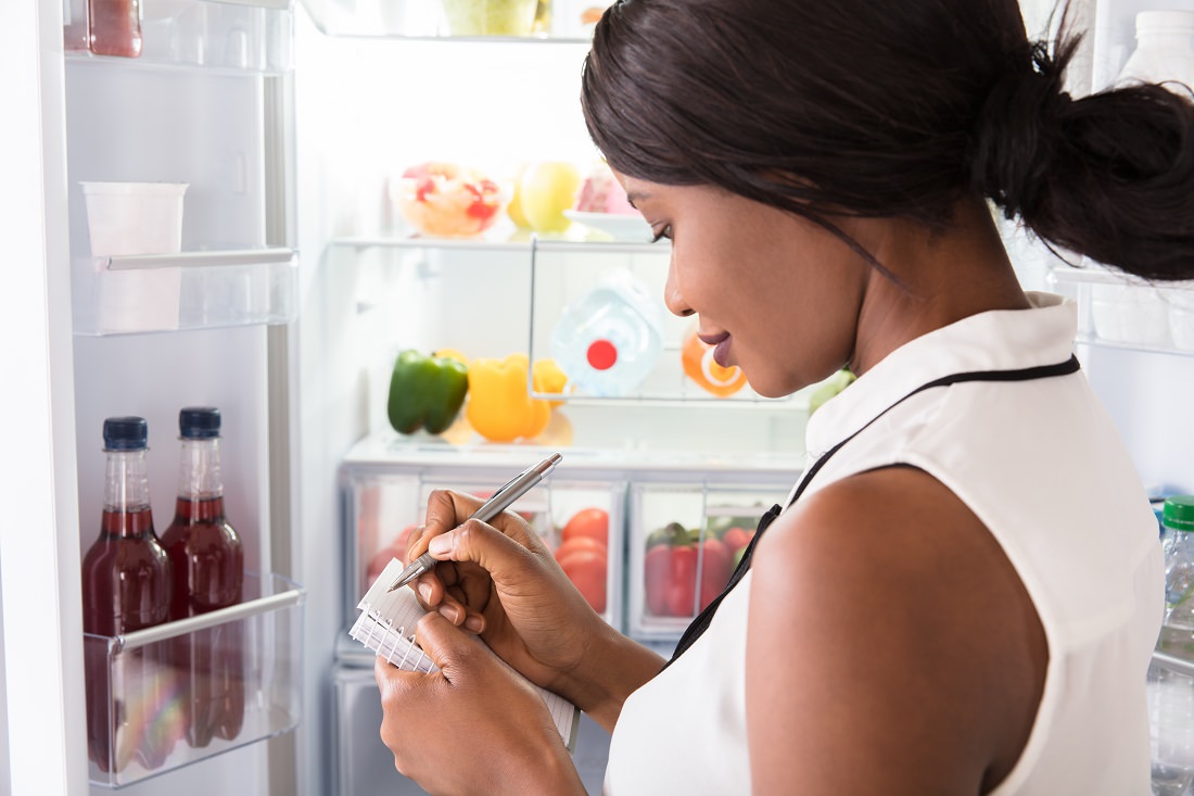 woman in front of open refrigerator, writing shopping list