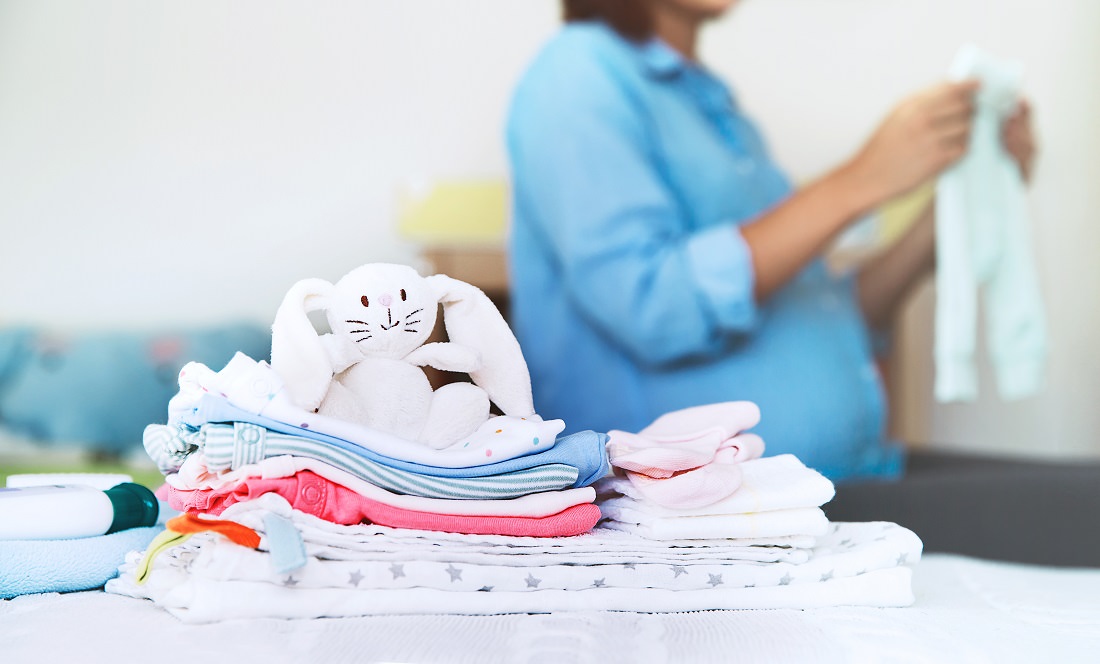 Pile of baby clothes on bed with pregnant woman in background