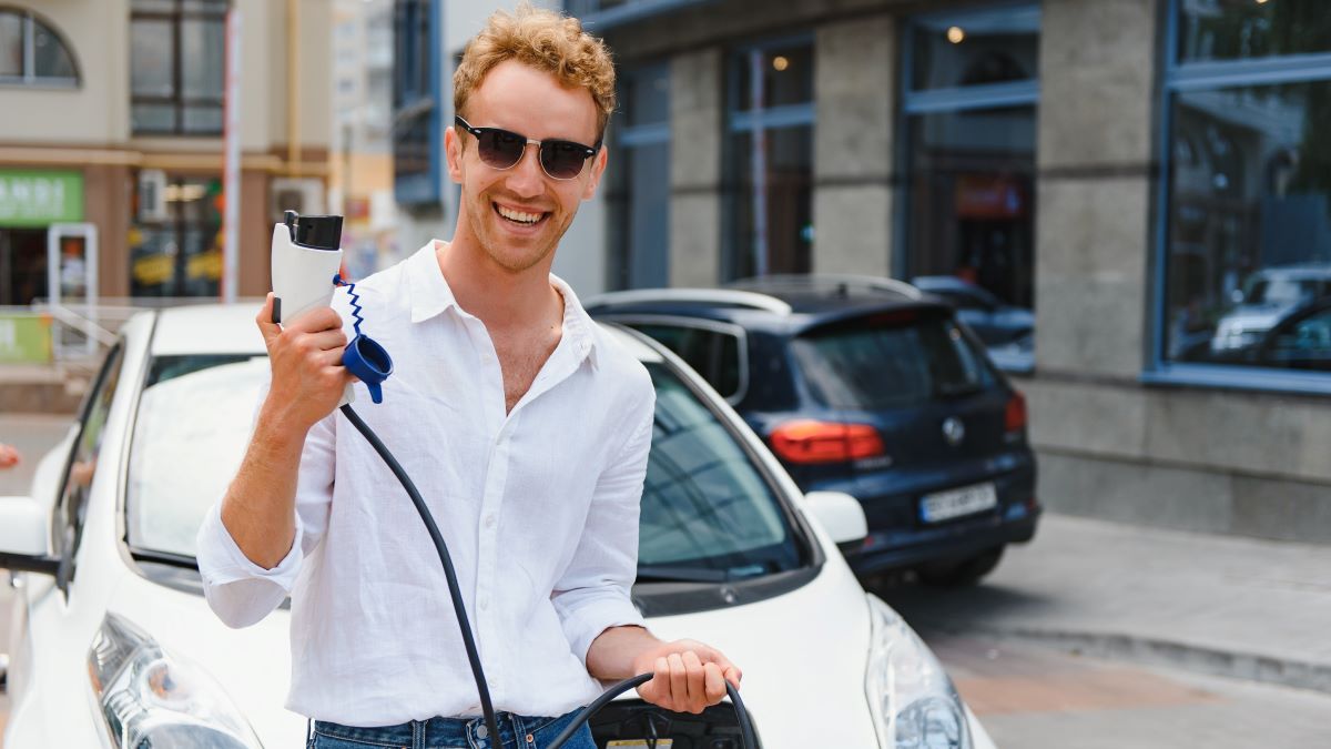 Smiling man standing by electric car holding charging cable