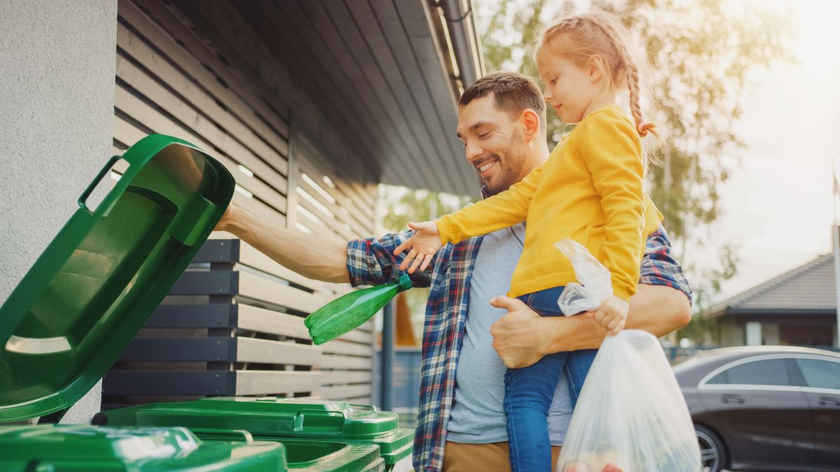 Father holds child as she throws bottle into recycling bin